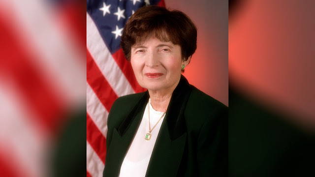 A formal portrait of retired Rear Adm. Faye Glenn Abdellah wearing a dark blazer and white blouse, standing in front of an American flag.